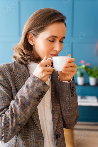 Beautiful woman with brown hair standing in the office and inhaling the aroma of fresh coffee.