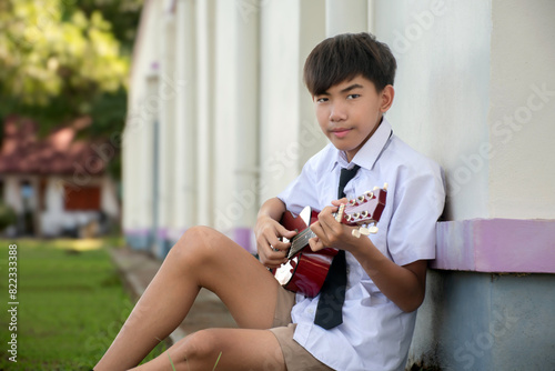 Asian preteen schoolboy in school uniform sitting near cream wall and playing ukulele after school.