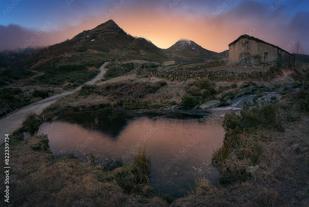 Naklejka premium warm sunrise at the Pardo waterfall, in the mountain pass of Estacas de Trueba, Burgos, with a small pond in the foreground and a pasiega cabin between mountains