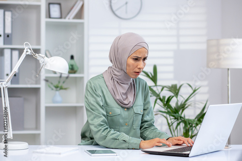 Frustrated woman in a hijab sitting at a desk while working on a laptop in a modern office setting.