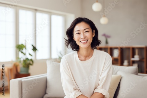 Portrait of a glad asian woman in her 40s smiling at the camera while standing against crisp minimalistic living room
