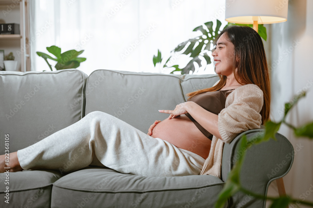 pregnancy, motherhood, anticipation, pregnant, parenting, stomach, parenthood, mother, touching, relaxing. A woman is sitting on a couch with a baby bump. She is smiling and she is relaxed. pregnancy, motherhood, anticipation, pregnant, parenting, stomach, parenthood, mother, touching, relaxing. A woman is sitting on a couch with a baby bump. She is smiling and she is relaxed.