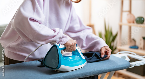 housekeeping, hygiene, laundry, domestic, garment, housewife, housework, maid, steam, clothes. A woman is ironing a shirt on an ironing board. The iron is hot and she is holding it with both hands.