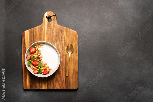 Fresh Greek Yogurt with granola and strawberries decorated with aromatic mint leaves on black stone background top view, copy space for your design. Healthy breakfast concept.