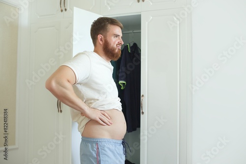 Unhappy sad upset young man with fat on her stomach, guy worried about body fat touching belly standing at home near closet