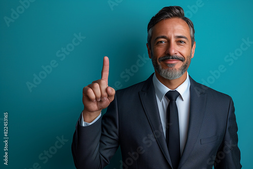 A handsome middle-aged man in a business suit, isolated on an empty blue background, with a smile, positive and confident expression, pointing his finger up, showing an idea, success concept 