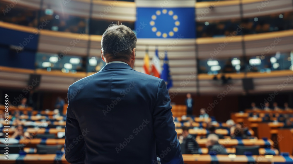 A man stands in front of a large audience in a room with a flag behind him A man stands in front of a large audience in a room with a flag behind him