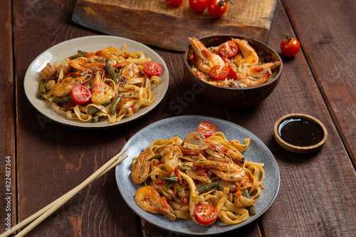 Rice with shrimp and udon in yaki soba sauce on a wooden table next to cherry tomatoes, soy sauce and chopsticks