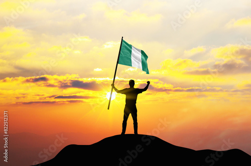 Nigeria flag being waved by a man celebrating success at the top of a mountain against sunset or sunrise. Nigeria flag for Independence Day.