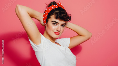 A young girl with dark brown hair in a red polka dot headband flexes her arm against a pink background, her white tee and cheeky smile radiating sass and strength. photo