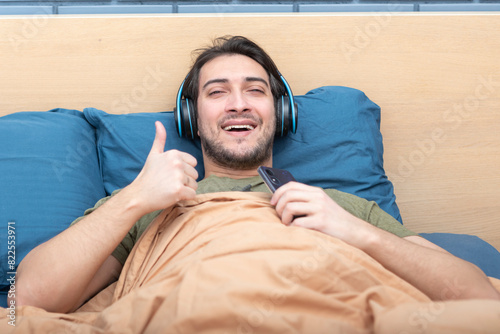 Young man lying in bed and listening to music using wireless headphones 