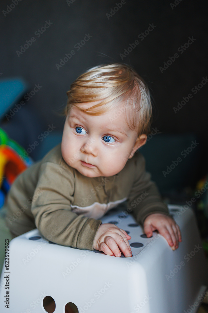 Fototapeta premium A small baby boy with blue eyes and blond hair is standing near a chair. A cute emotional baby is learning to stand on his feet
