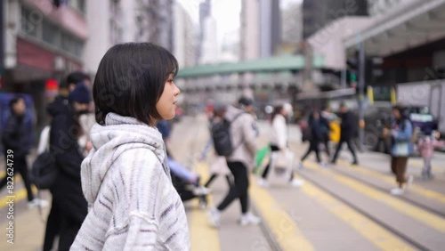 In Hong Kong's Causeway Bay, a young woman in her twenties navigates a busy crosswalk amidst bustling city life. The dynamic urban scene is filled with people and traffic. photo