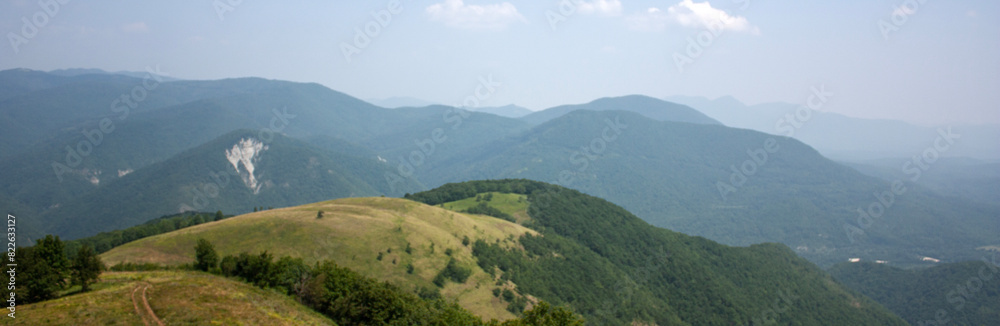 View of the road descending from the top of the mountain. On the horizon are mountains covered with green forest on a summer day. Clouds in the blue sky over green mountains on a sunny day. View of the road descending from the top of the mountain. On the horizon are mountains covered with green forest on a summer day. Clouds in the blue sky over green mountains on a sunny day.