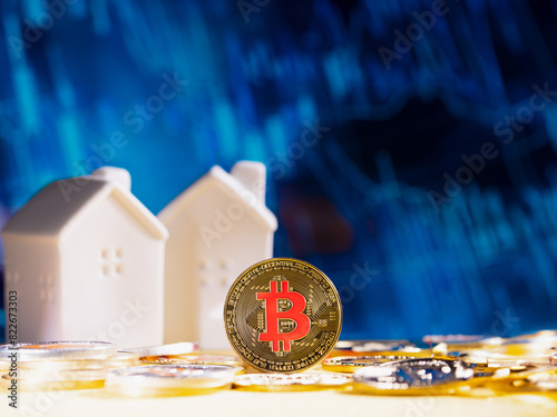 Bitcoin coin stands against a blurred blue background with financial graphs and two white model houses in the background. cryptocurrency and real estate. finance and investment on stock market