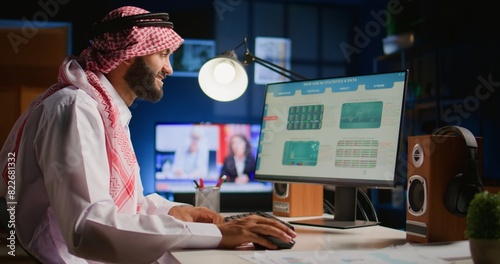 Cheerful muslim broker investor comparing stock exchange valuation financial profit numbers. Arab stockholder in dimly illuminated room looking at market shares growth charts, taking notes photo