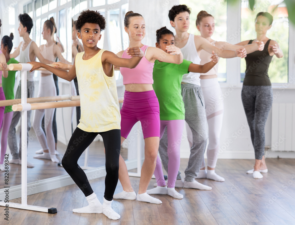 Thin teenage boy practicing plie position of ballet holding by ballet barre during choreography classes