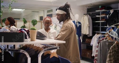 Customer receiving advice from employee in clothing store during promotional season while looking to buy cheap garments to fill his wardrobe. Man helped by worker to find sales in fashion boutique