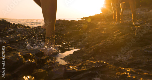 LENS FLARE  CLOSE UP  Barefoot woman walks with her dog on a rocky beach at sunset. Rugged Adriatic seashore glows in golden sunlight during their evening stroll. Active summer holidays at seaside.