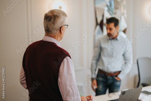 Two businessmen engage in a discussion at a bright office environment, with the focus on the senior man.