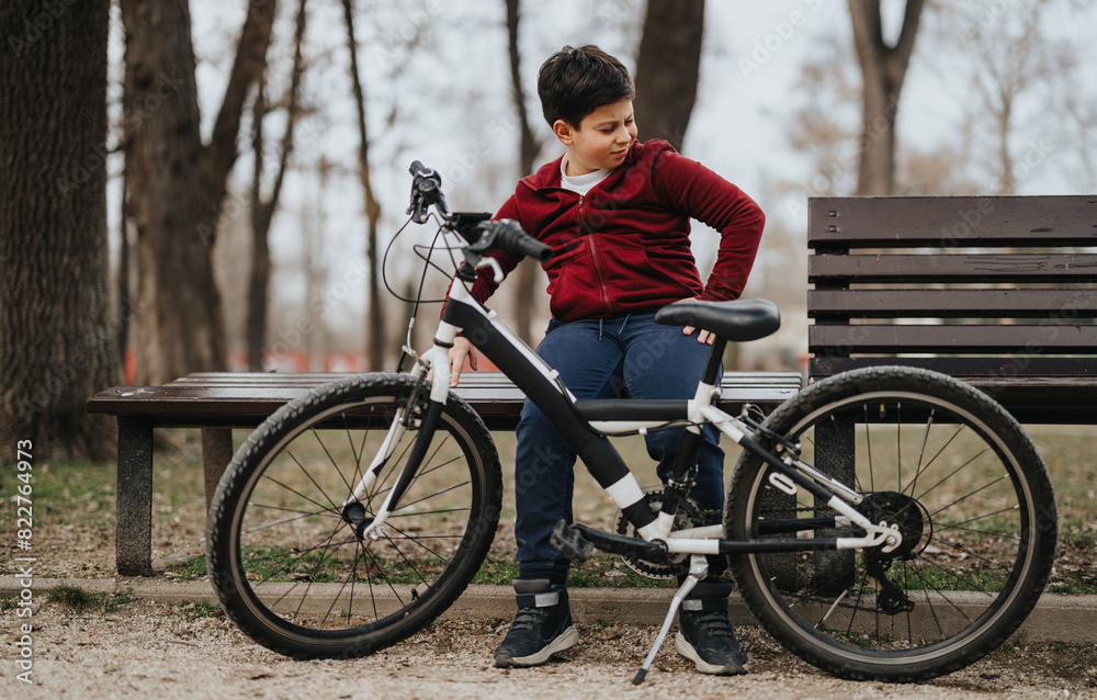 Fototapeta premium A young boy takes a break on a park bench beside his bicycle, enjoying a moment of leisure outdoors.