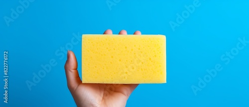 Yellow cleaning sponge gripped firmly in a human hand, symbolizing household chores and hygiene maintenance, with ample copy space on a striking bright blue background