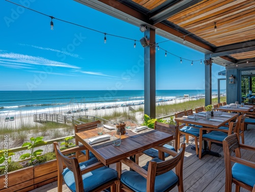 A beachside restaurant with tables and chairs overlooking the ocean. The tables are set with silverware and glasses, and the chairs are blue. Scene is relaxed and inviting