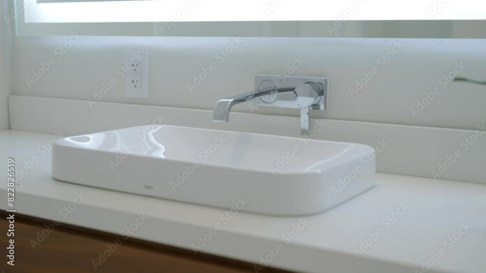 Rectangular bathroom sink with sleek faucet mounted on wall above it. White countertop and large window behind it allowing natural light to illuminating space Rectangular bathroom sink with sleek faucet mounted on wall above it. White countertop and large window behind it allowing natural light to illuminating space