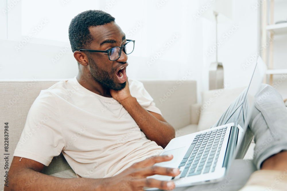 Smiling African American Freelancer Working on Laptop in Modern Home Office The young black man is sitting on a cozy sofa, typing on his computer with a focused expression Surrounded by the comforts