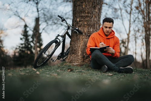 A young adult male in an orange hoodie rests against a tree with his bike beside him, writing something in his notebook. © qunica.com