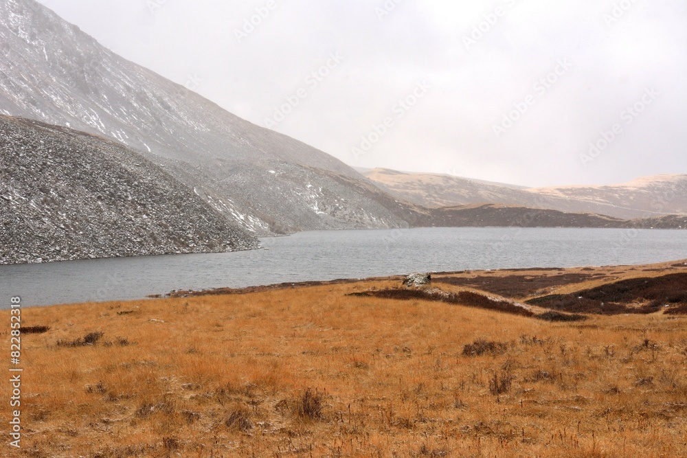 A fragment of a large lake with rocky shores overgrown with yellowed grass at the foot of a sheer cliff sprinkled with the first snow.