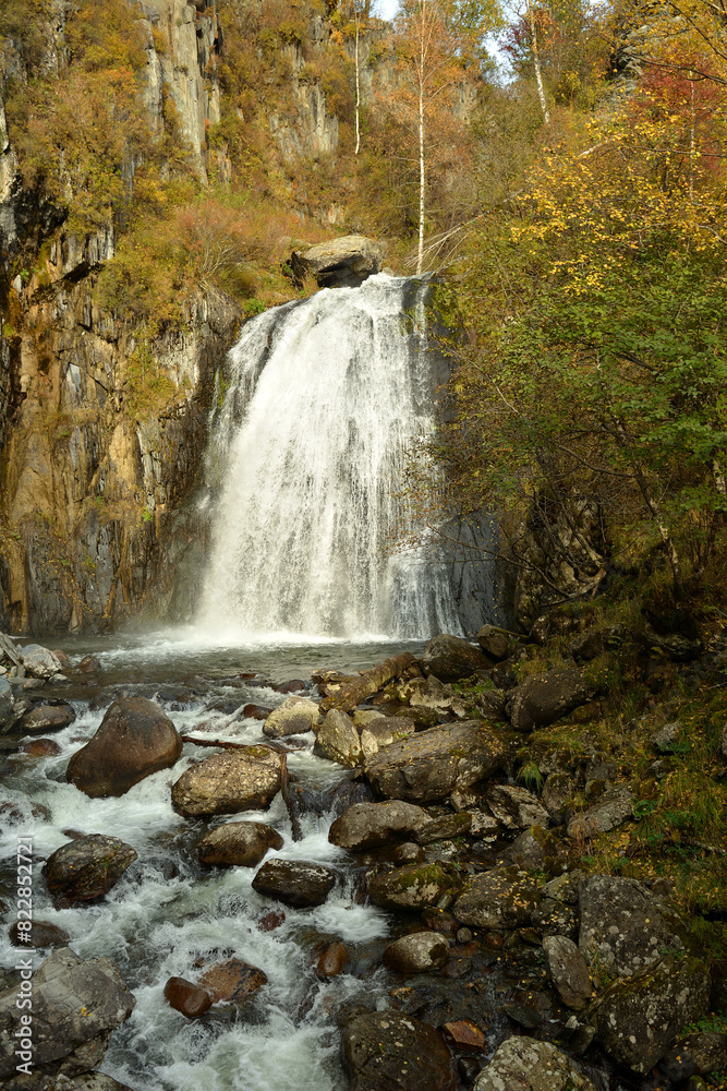 A stormy river flows like a rapid waterfall from a high mountain into an autumn forest on a cloudy day.