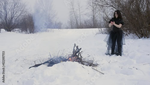 Woman brunette witch near bonfire in snowy woodland washes hands by snow in black dress. Ancient mystical spiritual dancing ceremony in winter forest. Scary wizard making ordinance standing on snow.