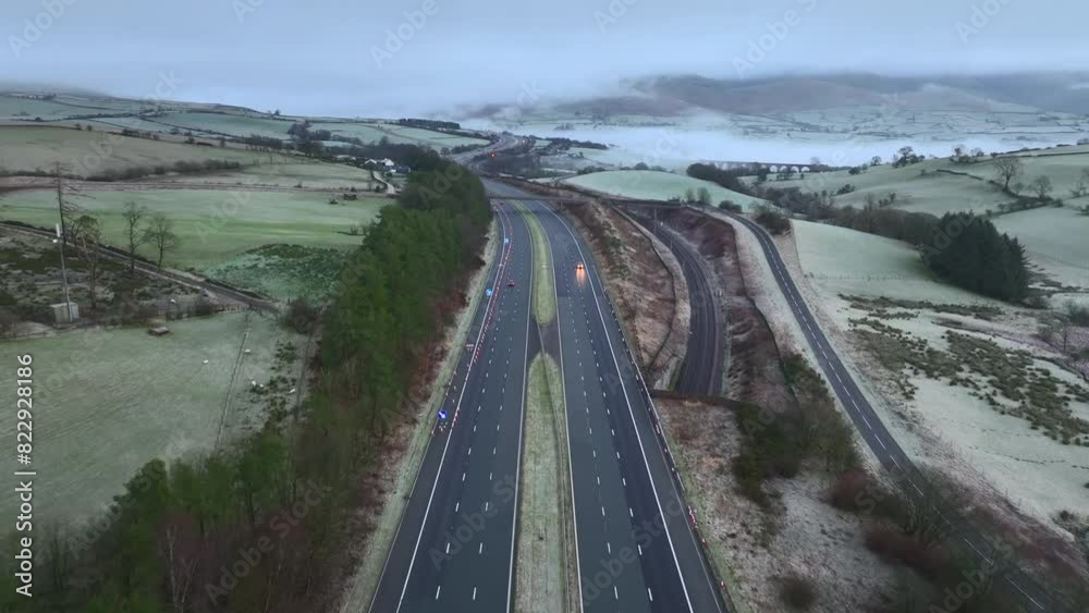 Flying down the centre of M6 highway with light traffic at dawn in winter with background hills covered in mist, fog and low cloud. Tebay, English Lake District, Cumbria, UK. Flying down the centre of M6 highway with light traffic at dawn in winter with background hills covered in mist, fog and low cloud. Tebay, English Lake District, Cumbria, UK.