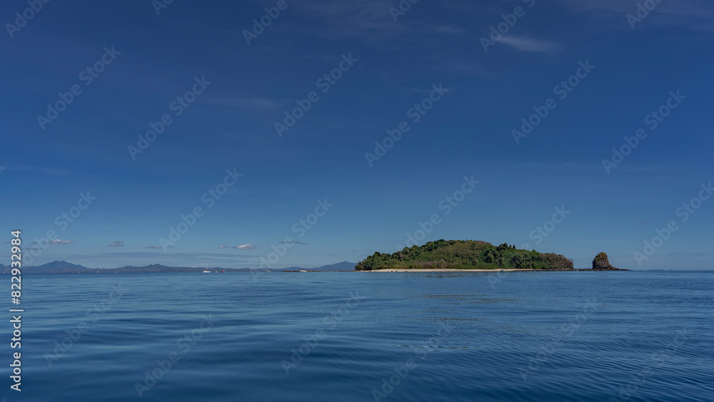 A beautiful tropical island in the blue ocean is covered with lush green vegetation. There are tiny silhouettes of people on the sandy beach. The boats are moored near the shore. The shiny water. 