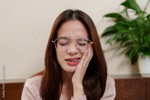 Canvas Print Young Asian girl with braces frowning, holding cheek in pain, wearing glasses and pink sweater