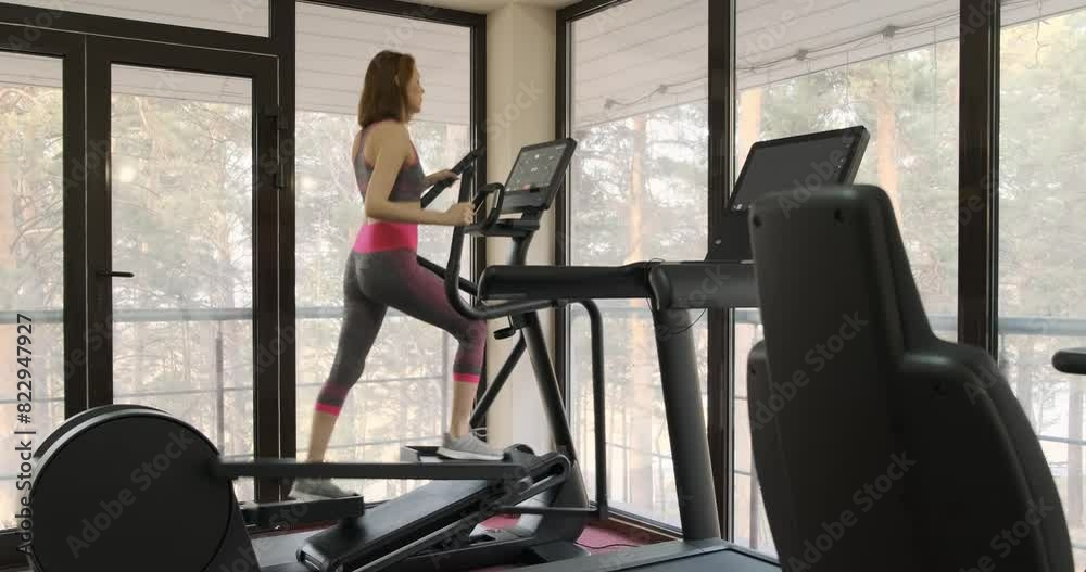 Middle aged woman working on stepper and looking at fitness bracelet while checking her activity and heart rate, gym workout. Girl is working out at gym. Young woman in sportswear out at sport hall Middle aged woman working on stepper and looking at fitness bracelet while checking her activity and heart rate, gym workout. Girl is working out at gym. Young woman in sportswear out at sport hall