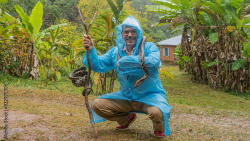 A man in a raincoat squatted down, smiling happily. Snakes wrapped around his neck and stick in hand.  Shiny patterned skin is visible, the boa constrictor's tongue is sticking out.  Madagascar. 
