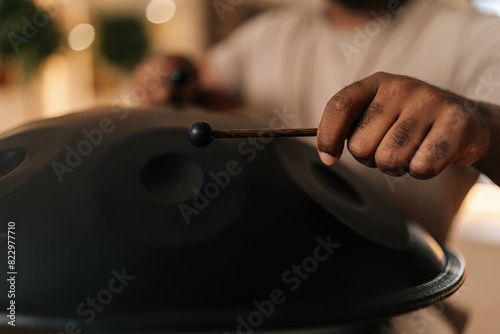 Close-up hands of unrecognizable black male in trance playing meditative instrument tank drum using sticks sitting in meditation room. Concept of mindfulness, tranquility, harmony, sound therapy.