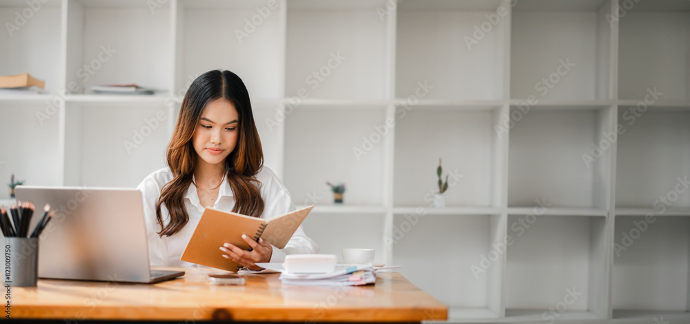 A woman is sitting at a desk with a laptop and a book. She is reading the book while looking at the laptop. The room is clean and organized, with a few potted plants and a vase on the table A woman is sitting at a desk with a laptop and a book. She is reading the book while looking at the laptop. The room is clean and organized, with a few potted plants and a vase on the table