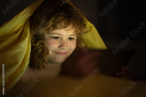 Kid alone at home. Kid playing game on tablet in bed at night. Kids with social media. Child lying in bed playing a tablet in dark room, light under blacket. Closeup kid watching cartoons on tablet. photo