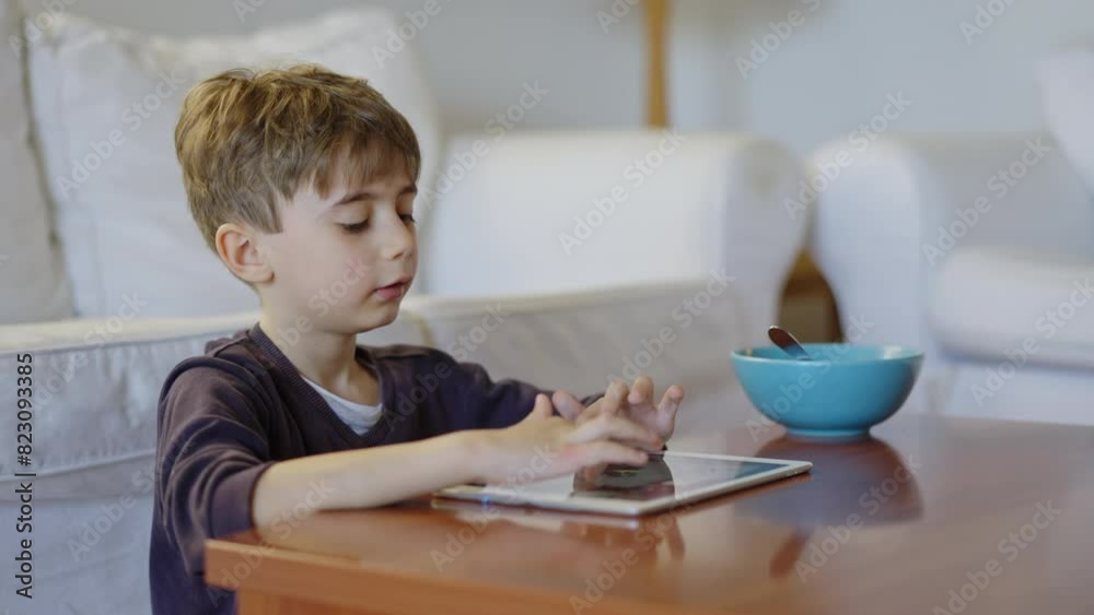 A boy engages with a digital tablet while eating breakfast from a bowl, seated in a cozy living room. A boy engages with a digital tablet while eating breakfast from a bowl, seated in a cozy living room.