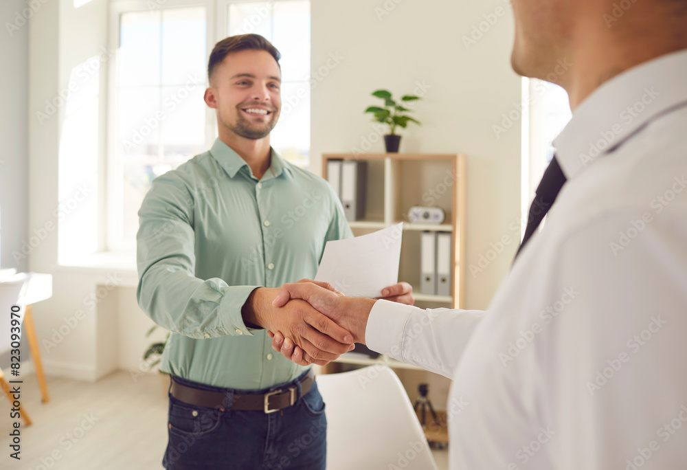 Obraz premium Two happy young men shaking hands and smiling. Two people meet in the office, establish partnership relations, make a lucrative business deal and exchange handshakes. Cooperation, teamwork concept