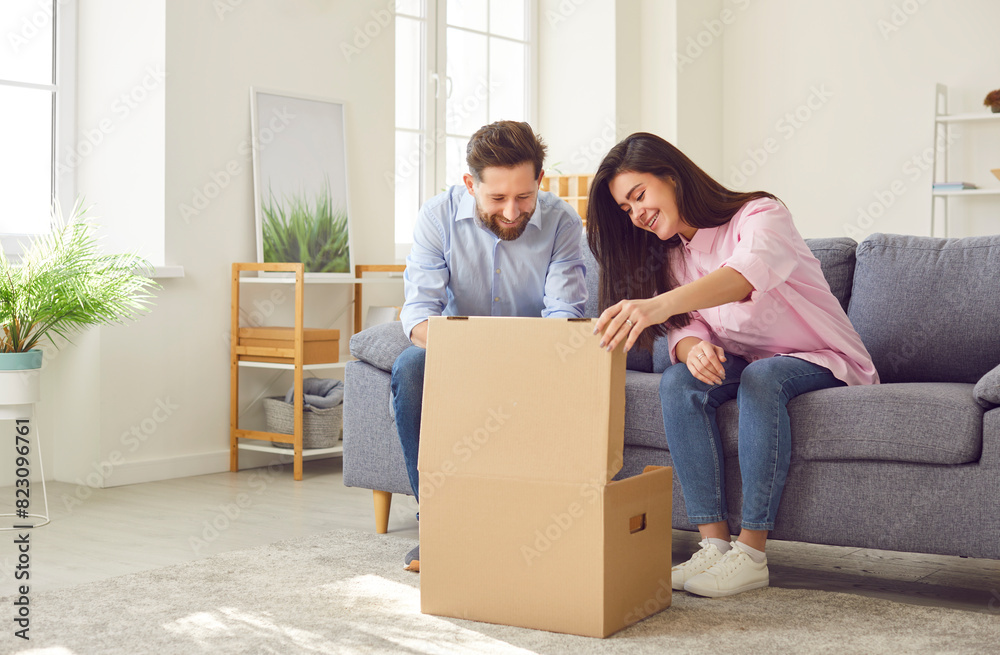 Happy young joyful couple unpacking parcel at home, sitting in the living room on sofa and smiling looking into open cardboard box. Delivery, online shopping, receive the parcel concept.