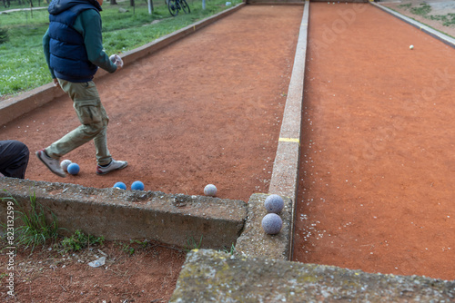 A player is walking on a red boules court, ready to throw blue boules. The court is bordered by concrete and stone, set in a park. The image captures the focus and concentration of the game.