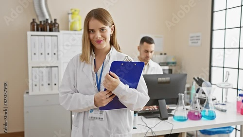 Wallpaper Mural A smiling woman scientist with a clipboard stands in a laboratory while a male colleague works in the background. Torontodigital.ca