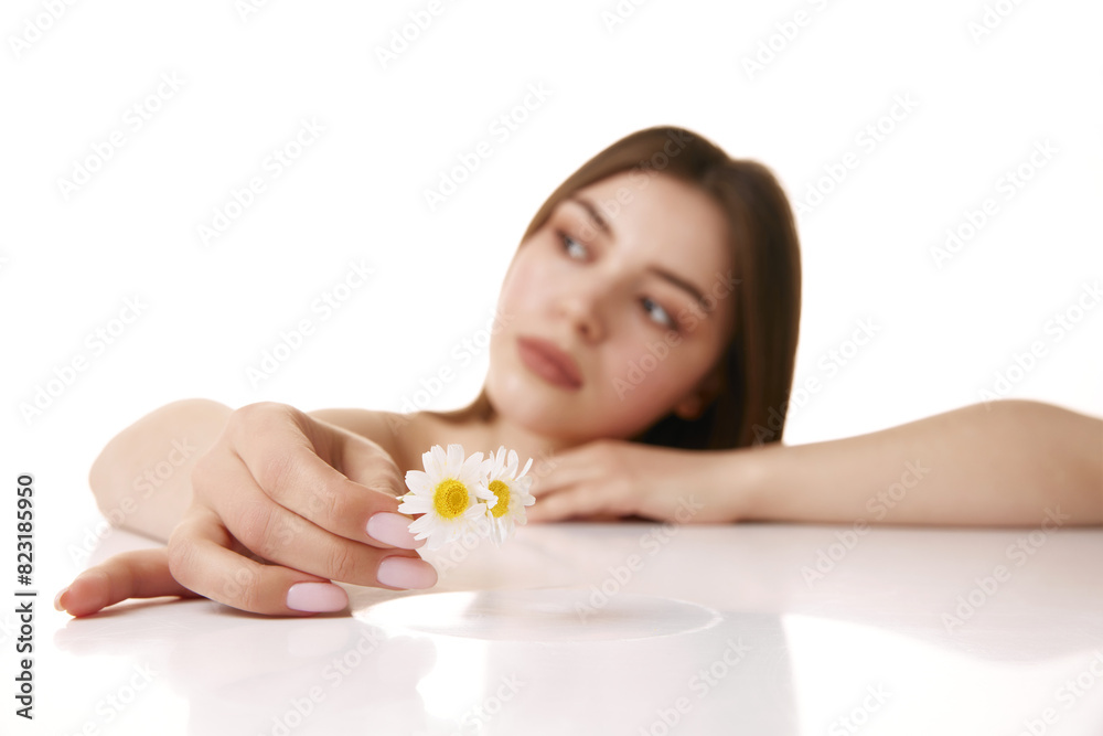 Young, beautiful woman with gentle expression, holding daisy looking away against white studio background. Selective focus. Concept of natural beauty and appearance, spa procedures and facial care. Young, beautiful woman with gentle expression, holding daisy looking away against white studio background. Selective focus. Concept of natural beauty and appearance, spa procedures and facial care.