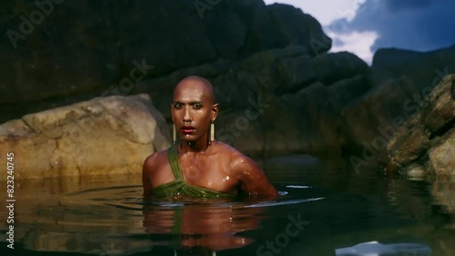 Androgynous person of color dives out of water in boutique open dress on rocky ocean backwater in still water inside a creek on coastline. Trans sexual fashion model in skimpy outfit wears jewellery.