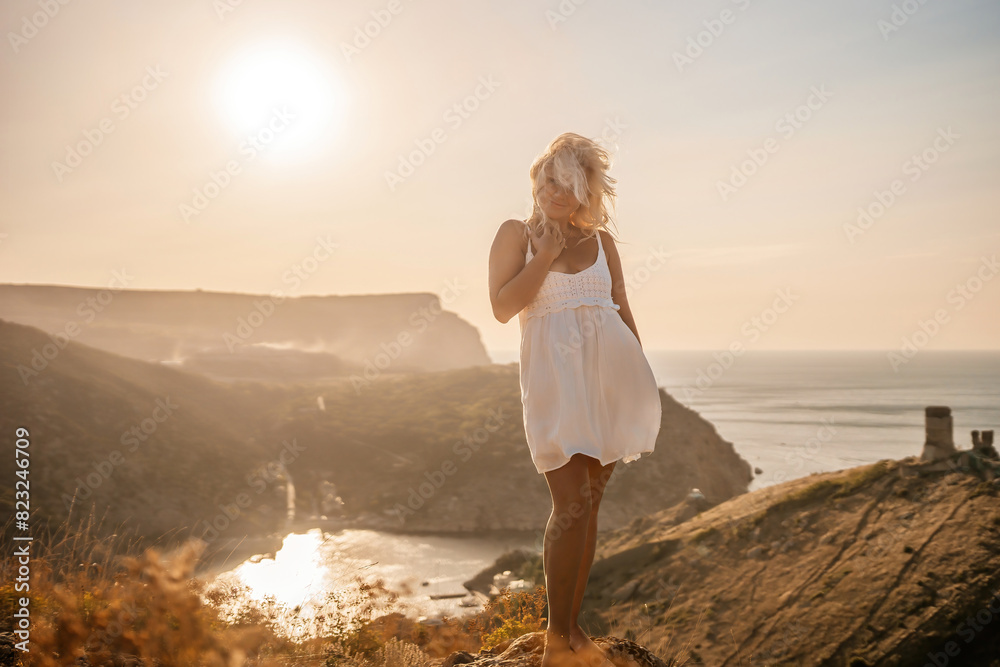 A blonde woman stands on a hill overlooking the ocean. She is wearing a white dress and she is enjoying the view.