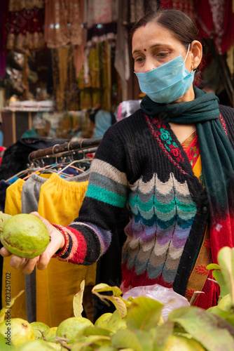 Indian mature woman wearing protective face mask and buying guava from outdoor market 